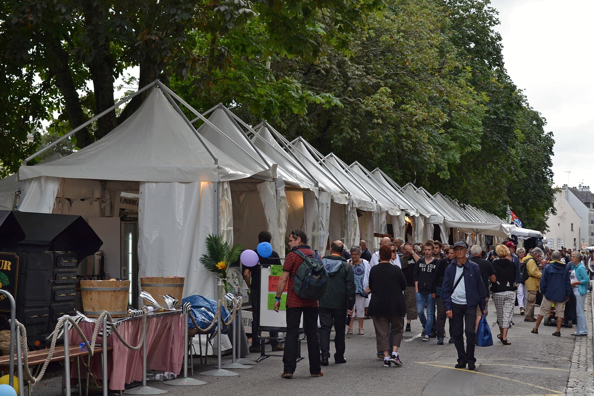 Une rangée de barnums blancs le long d'une rue ombragée, pendant une fête de ville ; les visiteurs passent entre les stands.